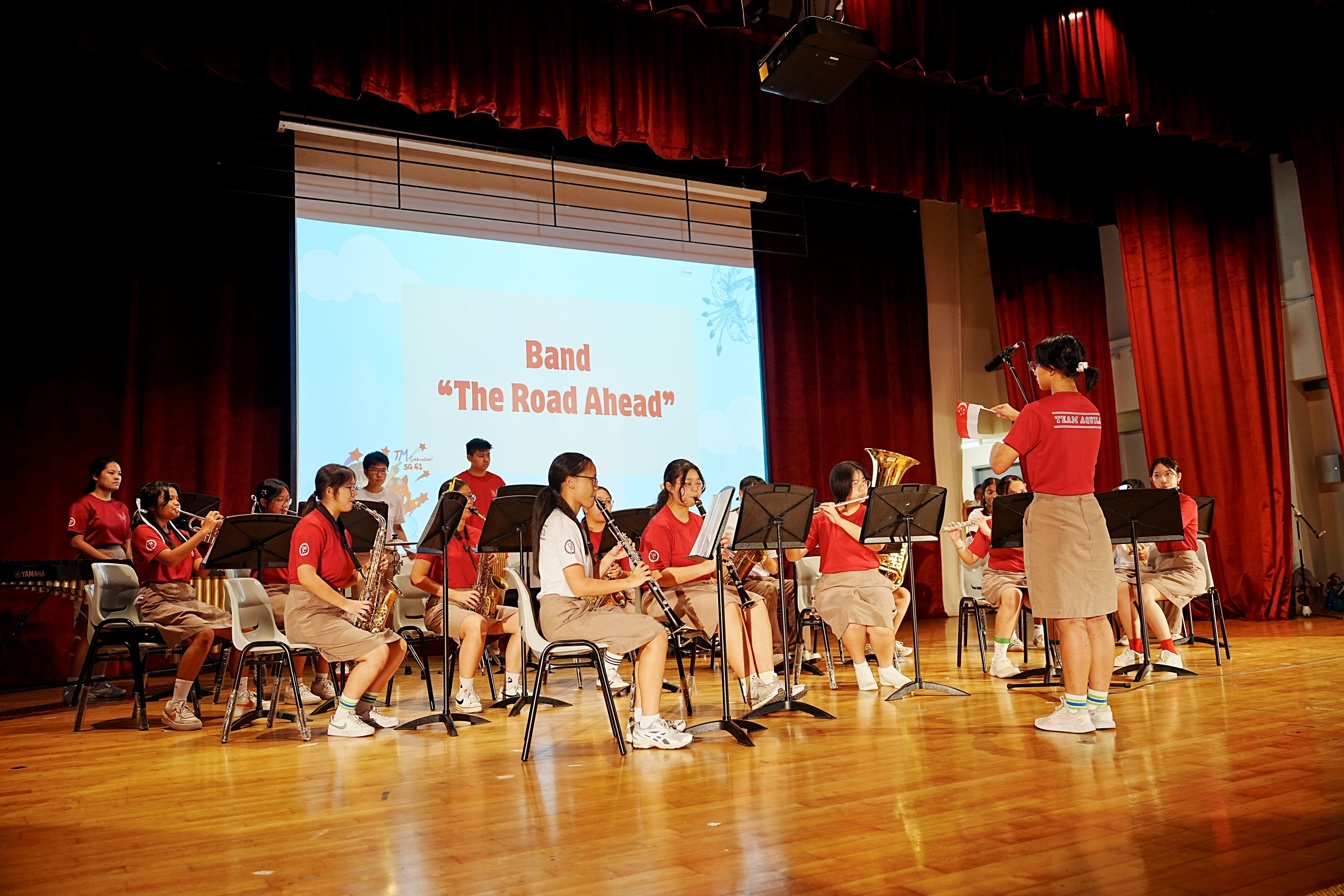 Students in red shirts perform on stage under a projected slide reading "Band – The Road Ahead", led by a conductor.
